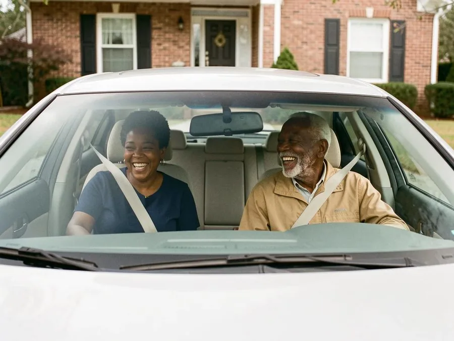 A caregiver and a laughing older man buckled into the front seats of a car parked outside a brick house